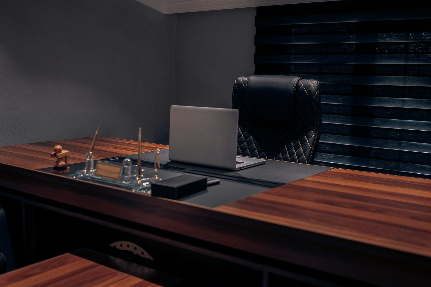 Bureau exécutif en bois sombre dans une pièce tamisée, fauteuil en cuir matelassé, ordinateur portable fermé, encrier et porte-stylos en laiton, atmosphère feutrée.