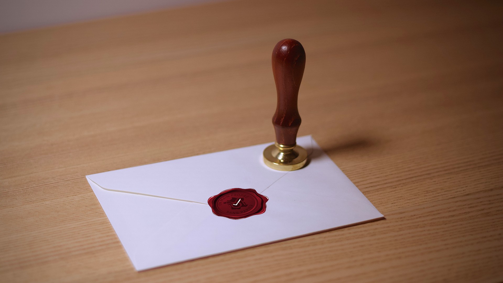 Enveloppe blanche scellée par un cachet de cire rouge marqué « Remis. En main propre. Sans papier. », tampon à manche en bois et laiton posé derrière, sur un plan de travail en bois clair, vue de trois quarts.